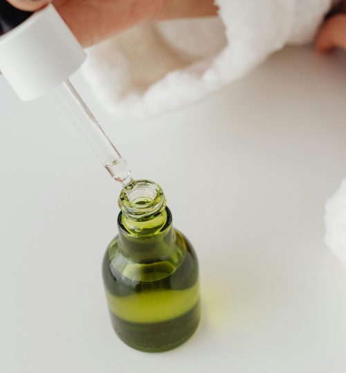 Close-up of hands with manicured nails holding a dropper over a green serum bottle.